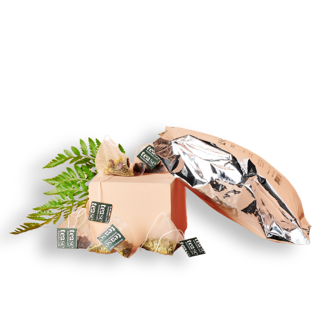 Tease Wellness Salon & Spa Refills with assorted herbal tea bags and open metallic pouch next to green fern leaves.